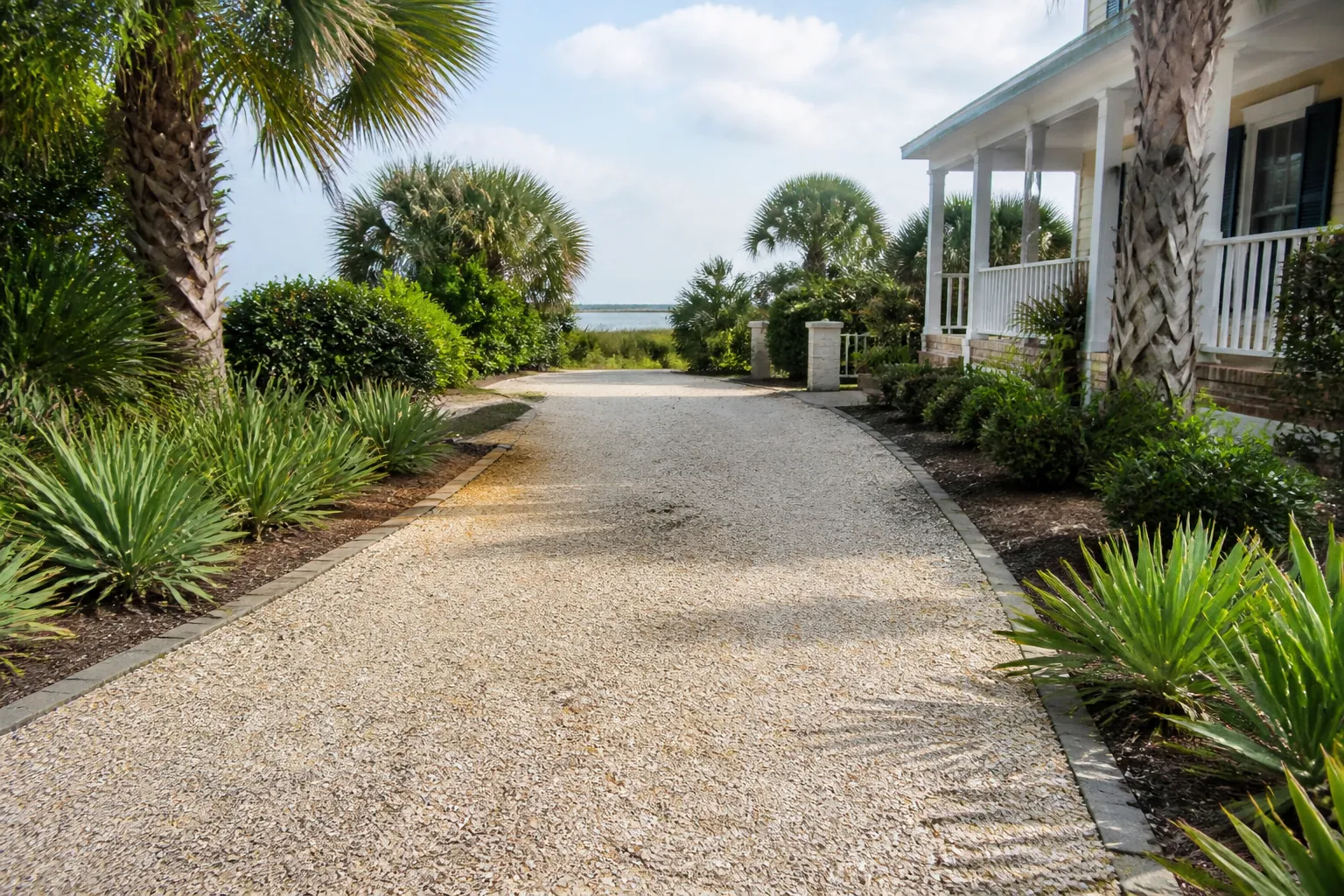 Shell Driveway Beside a Palm-Lined Coastal Home A pale shell driveway bordered by pavers, palms and low greenery beside a white porch, with water visible in the distance.